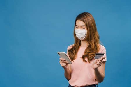 Young Asia lady wearing medical face mask using phone and credit bank card with positive expression, smiles broadly, dressed in casual clothing and stand isolated on blue background.の写真素材