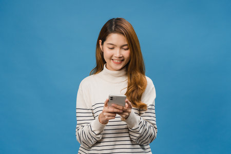 Young Asia lady using phone with positive expression, smiles broadly, dressed in casual clothing feeling happiness and standing isolated on blue background. Happy adorable glad woman rejoices success.の写真素材