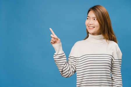 Portrait of young Asian lady smiling with cheerful expression, shows something amazing at blank space in casual clothing and standing isolated over blue background. Facial expression concept.の写真素材