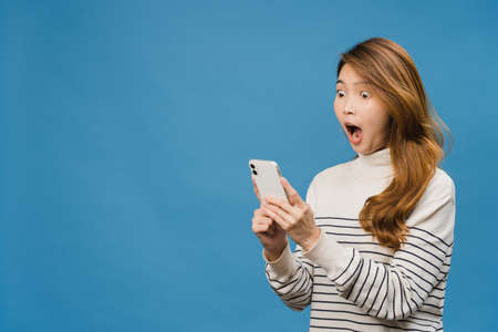 Surprised young Asia lady using mobile phone with positive expression, smiles broadly, dressed in casual clothing and standing isolated on blue background. Happy adorable glad woman rejoices success.の写真素材