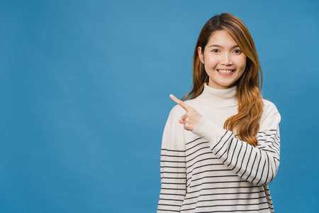 Portrait of young Asian lady smiling with cheerful expression, shows something amazing at blank space in casual clothing and looking at camera isolated over blue background. Facial expression concept.の写真素材