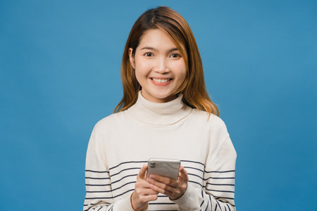 Surprised young Asia lady using mobile phone with positive expression, smile broadly, dressed in casual clothing and looking at camera on blue background. Happy adorable glad woman rejoices success.の写真素材