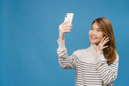 Smiling adorable Asian female making selfie photo on smart phone with positive expression in casual clothing and stand isolated on blue background. Happy adorable glad woman rejoices success.の写真素材