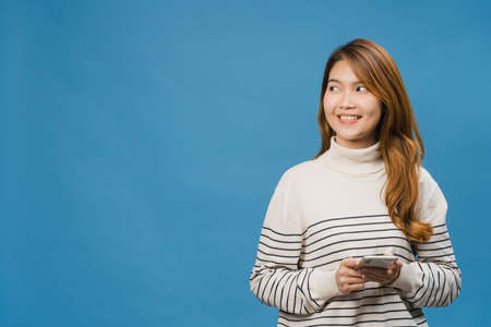 Young Asia lady using phone with positive expression, smiles broadly, dressed in casual clothing feeling happiness and standing isolated on blue background. Happy adorable glad woman rejoices success.の写真素材