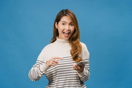 Surprised young Asia lady using mobile phone with positive expression, smiles broadly, dressed in casual clothing and looking at camera on blue background. Happy adorable glad woman rejoices success.の写真素材