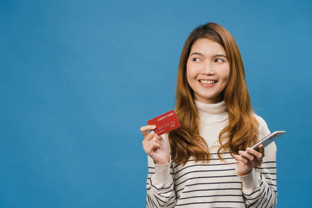 Young Asia lady using phone and credit bank card with positive expression, smiles broadly, dressed in casual clothing and stand isolated on blue background. Happy adorable glad woman rejoices success.の写真素材