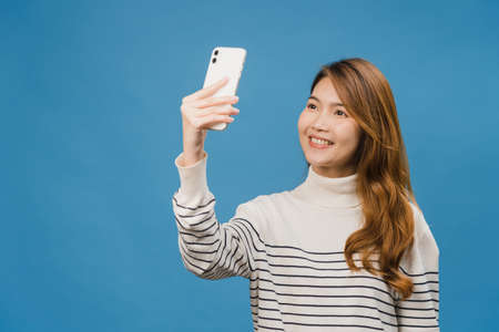 Smiling adorable Asian female making selfie photo on smart phone with positive expression in casual clothing and stand isolated on blue background. Happy adorable glad woman rejoices success.の写真素材