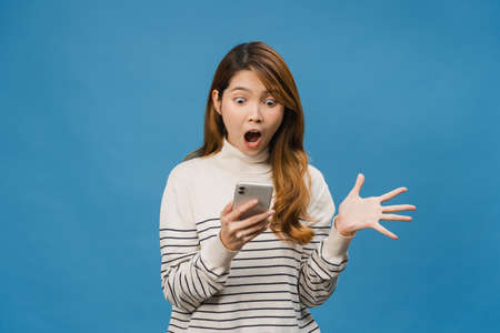 Surprised young Asia lady using mobile phone with positive expression, smiles broadly, dressed in casual clothing and standing isolated on blue background. Happy adorable glad woman rejoices success.の写真素材