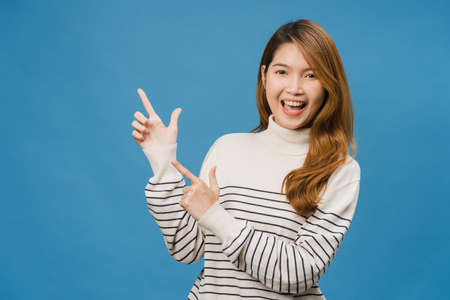 Portrait of young Asian lady smiling with cheerful expression, shows something amazing at blank space in casual clothing and looking at camera isolated over blue background. Facial expression concept.の写真素材