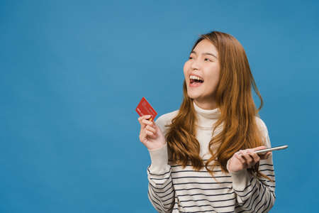 Young Asia lady using phone and credit bank card with positive expression, smiles broadly, dressed in casual clothing and stand isolated on blue background. Happy adorable glad woman rejoices success.の写真素材