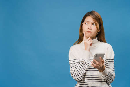 Thinking dreaming young Asia lady using phone with positive expression, dressed in casual clothing feeling happiness and stand isolated on blue background. Happy adorable glad woman rejoices success.の写真素材