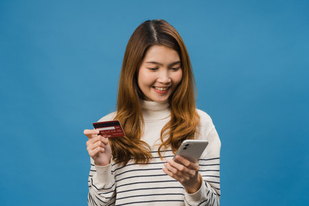 Young Asia lady using phone and credit bank card with positive expression, smiles broadly, dressed in casual clothing and stand isolated on blue background. Happy adorable glad woman rejoices success.の写真素材