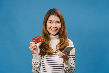 Young Asia lady using mobile phone and credit bank card with positive expression, dressed in casual cloth and looking at camera isolated on blue background. Happy adorable glad woman rejoices success.の写真素材