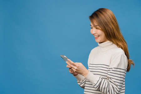 Young Asia lady using phone with positive expression, smiles broadly, dressed in casual clothing feeling happiness and standing isolated on blue background. Happy adorable glad woman rejoices success.の写真素材