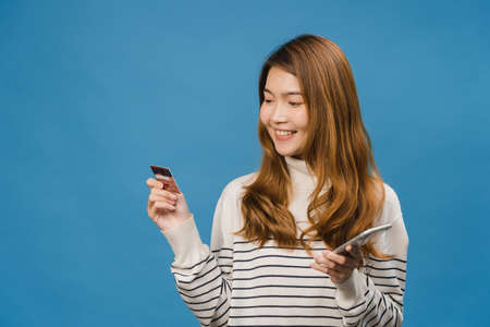 Young Asia lady using phone and credit bank card with positive expression, smiles broadly, dressed in casual clothing and stand isolated on blue background. Happy adorable glad woman rejoices success.の写真素材