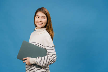 Young Asia lady hold laptop with positive expression, smiles broadly, dressed in casual clothing feeling happiness and stand isolated on blue background. Happy adorable glad woman rejoices success.の写真素材