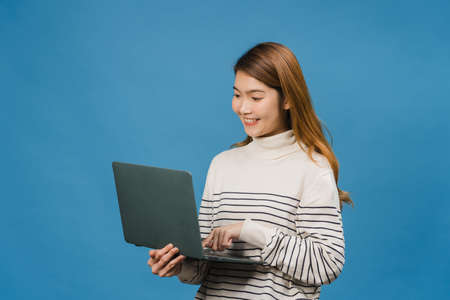 Young Asia lady using laptop with positive expression, smiles broadly, dressed in casual clothing feeling happiness and stand isolated on blue background. Happy adorable glad woman rejoices success.の写真素材