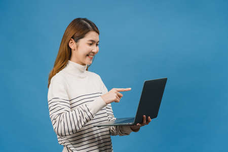 Young Asia lady using laptop with positive expression, smiles broadly, dressed in casual clothing feeling happiness and stand isolated on blue background. Happy adorable glad woman rejoices success.の写真素材