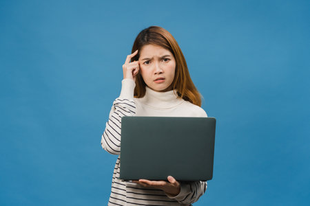 Young Asia lady using laptop with negative expression, excited screaming, cry emotional angry in casual cloth and stand isolated on blue background with blank copy space. Facial expression concept.の写真素材
