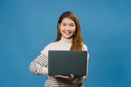 Surprised young Asia lady using laptop with positive expression, smile broadly, dressed in casual clothing and looking at camera on blue background. Happy adorable glad woman rejoices success.の写真素材