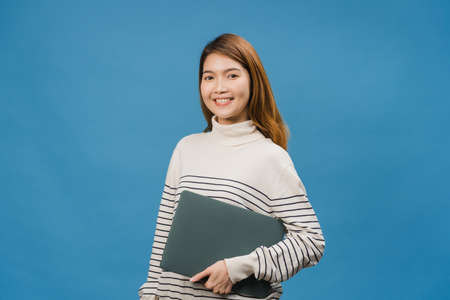 Surprised young Asia lady hold laptop with positive expression, smile broadly, dressed in casual clothing and looking at camera on blue background. Happy adorable glad woman rejoices success.の写真素材