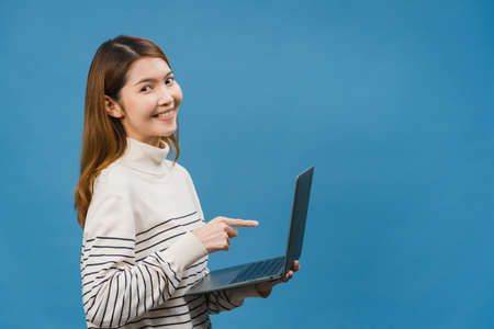 Young Asia lady using laptop with positive expression, smiles broadly, dressed in casual clothing feeling happiness and stand isolated on blue background. Happy adorable glad woman rejoices success.の写真素材