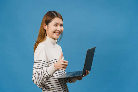 Young Asia lady using laptop with positive expression, smiles broadly, dressed in casual clothing feeling happiness and stand isolated on blue background. Happy adorable glad woman rejoices success.の写真素材