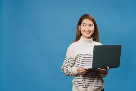 Young Asia lady using laptop with positive expression, smiles broadly, dressed in casual clothing feeling happiness and stand isolated on blue background. Happy adorable glad woman rejoices success.の写真素材