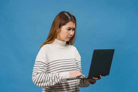 Young Asia lady using laptop with negative expression, excited screaming, cry emotional angry in casual cloth and stand isolated on blue background with blank copy space. Facial expression concept.の写真素材