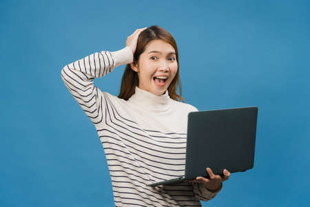 Young Asia lady using laptop with positive expression, smiles broadly, dressed in casual clothing feeling happiness and stand isolated on blue background. Happy adorable glad woman rejoices success.の写真素材