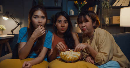Group of attractive Asia lady girl freaking out fear and terrified moment eat popcorn watch horror online movie on couch in living room at home in night. Weekend lifestyle activity quarantine concept.の写真素材