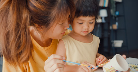 Happy cheerful Asia family mom teach toddler girl paint ceramic pot having fun relax on table in living room at house. Spending time together, Social distance, Quarantine for coronavirus prevention.の写真素材