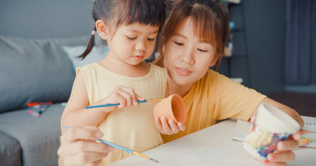Happy cheerful Asia family mom teach toddler girl paint ceramic pot having fun relax on table in living room at house. Spending time together, Social distance, Quarantine for coronavirus prevention.の写真素材