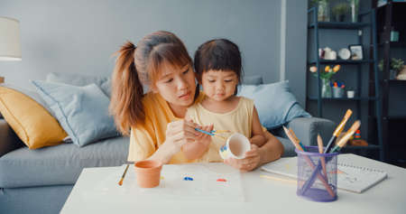 Happy cheerful Asia family mom teach toddler girl paint ceramic pot having fun relax on table in living room at house. Spending time together, Social distance, Quarantine for coronavirus prevention.の写真素材
