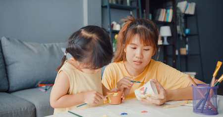 Happy cheerful Asia family mom teach toddler girl paint ceramic pot having fun relax on table in living room at house. Spending time together, Social distance, Quarantine for coronavirus prevention.の写真素材