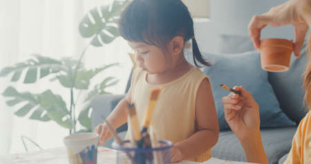 Happy cheerful Asia family mom teach girl paint use album and colourful pencils having fun relax on couch in living room at house. Spending time together, Social distance, Quarantine for coronavirus.の写真素材