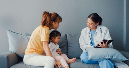 Young Asia female pediatrician hold stethoscope exam little girl patient visit doctor with mother sit on couch in living room at house. Medical care insurance, Visit patient at home concept.の写真素材