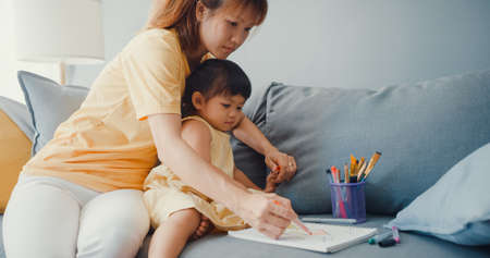 Happy cheerful Asia family mom teach girl paint use album and colourful pencils having fun relax on couch in living room at house. Spending time together, Social distance, Quarantine for coronavirus.の写真素材