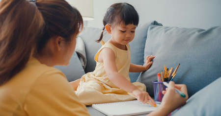 Happy cheerful Asia family mom teach girl paint use album and colourful pencils having fun relax on couch in living room at house. Spending time together, Social distance, Quarantine for coronavirus.の写真素材