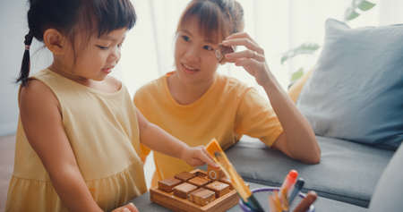 Happy cheerful Asia family mom teach girl paint use album and colourful pencils having fun relax on couch in living room at house. Spending time together, Social distance, Quarantine for coronavirus.の写真素材