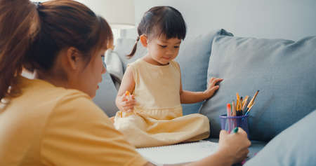 Happy cheerful Asia family mom teach girl paint use album and colourful pencils having fun relax on couch in living room at house. Spending time together, Social distance, Quarantine for coronavirus.の写真素材