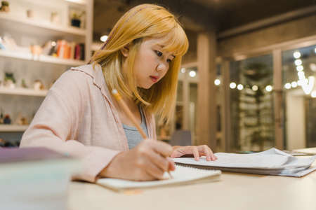 Asian student women reading books in library at university. Young undergraduate girl do homework, read textbook, study hard for knowledge on lecture desk at college campus overtime night.の写真素材