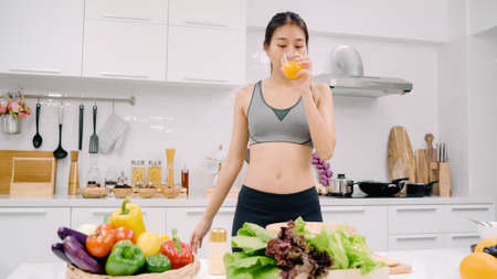 Young Asian woman drinking orange juice in the kitchen, beautiful female in sport clothing use organic fruits lots of nutrition making orange juice by herself at home. Healthy food concept.の写真素材