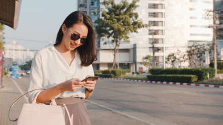 Successful young Asia businesswoman in fashion office clothes hailing on road catching taxi and holding smart phone while standing outdoors in urban modern city. Business on the go concept.の写真素材