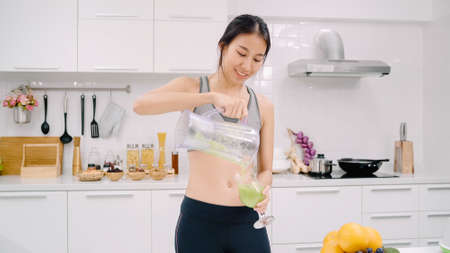 Young Asian woman drinking orange juice in the kitchen, beautiful female in sport clothing use organic fruits lots of nutrition making orange juice by herself at home. Healthy food concept.の写真素材