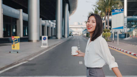 Successful young Asia businesswoman in fashion office clothes hailing on road catching taxi and holding smart phone while standing outdoors in urban modern city. Business on the go concept.の写真素材