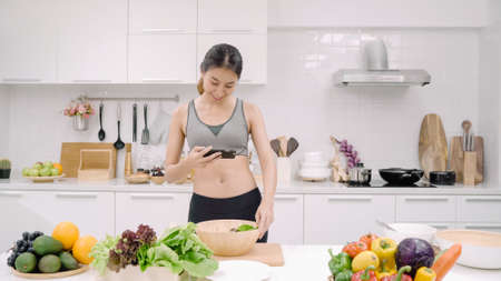 Happy Asian woman using tablet for looking recipe while making food in the kitchen, female use organic vegetable for healthy food at home. Lifestyle women making food concept.の写真素材