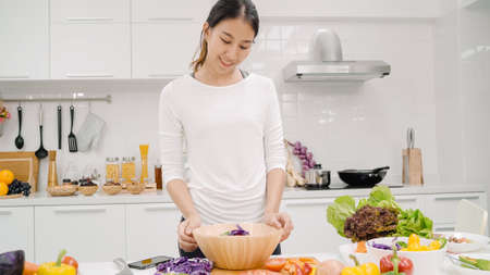 Happy Asian woman using tablet for looking recipe while making food in the kitchen, female use organic vegetable for healthy food at home. Lifestyle women making food concept.の写真素材