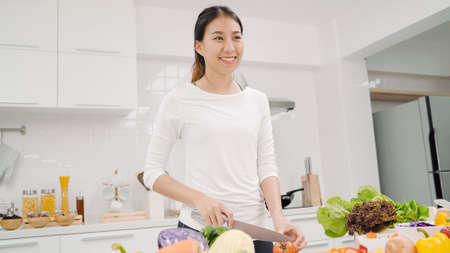 Happy Asian woman using tablet for looking recipe while making food in the kitchen, female use organic vegetable for healthy food at home. Lifestyle women making food concept.の写真素材