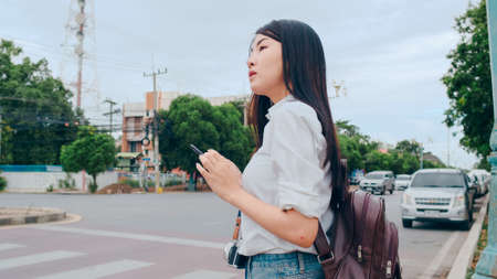 Young Asian backpacker woman tourist with camera stand in front of cross walk street look at mobile phone search navigation application for the location, Lifestyle tourist travel holiday concept.の写真素材